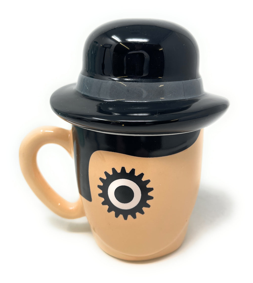 A Clockwork Orange Ceramic Mug shaped like a stylized face wearing a glossy black bowler hat, with a peach-beige body and a black circular eye graphic on the front, photographed on a white background.