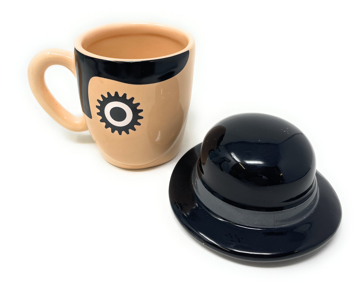 A Clockwork Orange Ceramic Mug shaped like a tan hat with a black band and a black gear emblem, shown next to a glossy black hat-shaped lid on a white background.