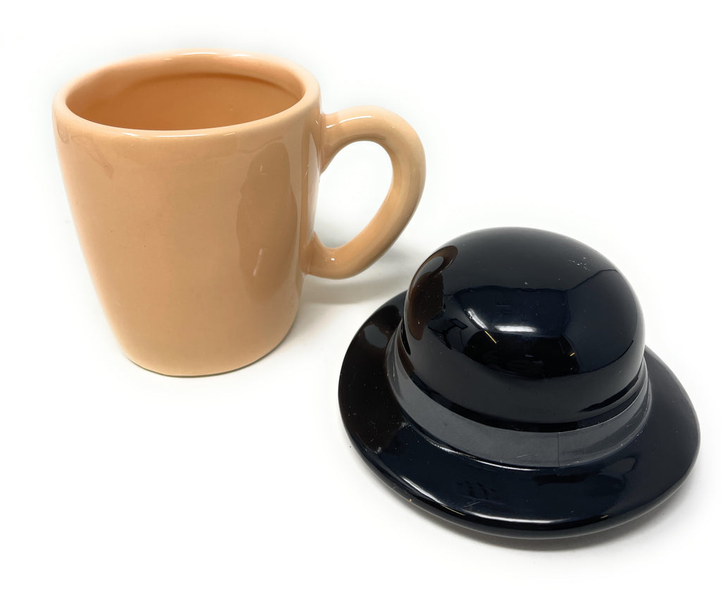A beige Clockwork Orange Ceramic Mug with a glossy finish next to a matching black ceramic lid shaped like a bowler hat, set against a plain white background.