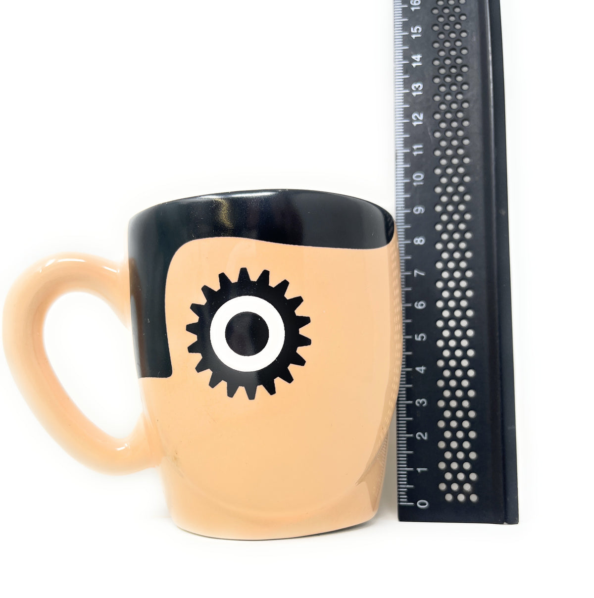 A peach-colored Clockwork Orange Ceramic Mug with a glossy black handle and rim, featuring a black gear graphic with a white center. A black ruler is positioned next to the mug for scale against a white background.