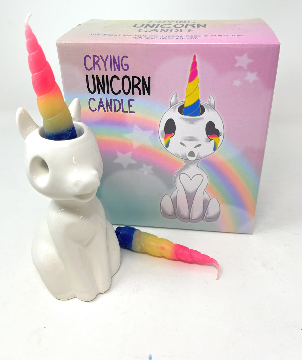 A ceramic unicorn-shaped candle in front of its pink packaging box, with a separate rainbow-colored wax horn protruding from the top of the unicorn head. The box features an illustration of a crying unicorn with a rainbow horn and the text 'CRYING UNICORN CANDLE'.