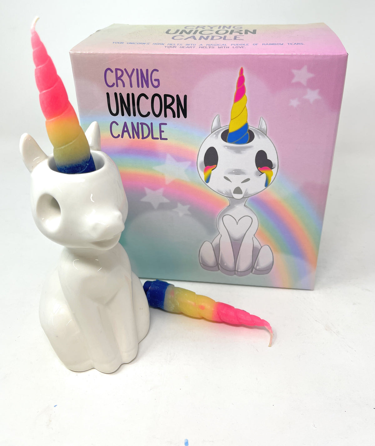 A ceramic unicorn-shaped candle in front of its pink packaging box, with a separate rainbow-colored wax horn protruding from the top of the unicorn head. The box features an illustration of a crying unicorn with a rainbow horn and the text 'CRYING UNICORN CANDLE'.
