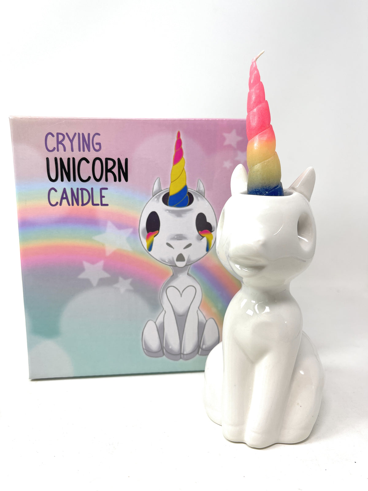 A white ceramic unicorn-shaped candle holder with a rainbow-colored horn, shown next to its pastel packaging box featuring a cartoon crying unicorn and the text 'CRYING UNICORN CANDLE'.