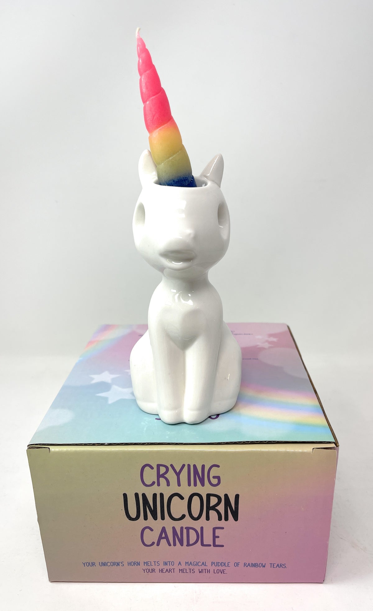A white ceramic unicorn-shaped candle holder with a rainbow-colored horn, displayed on top of its pastel-colored packaging box against a light gray background.