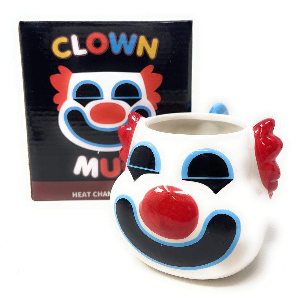 A white ceramic clown mug with a red nose, black and blue painted facial features, and red hair-like handles on the sides, shown in front of its black retail box featuring a clown face graphic and the word 'CLOWN'.