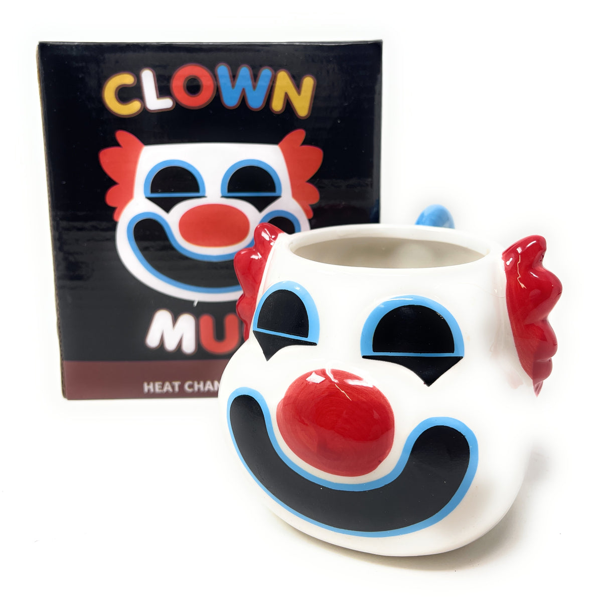 A white ceramic clown mug with a red nose, black and blue painted facial features, and red hair-like handles on the sides, shown in front of its black retail box featuring a clown face graphic and the word 'CLOWN'.