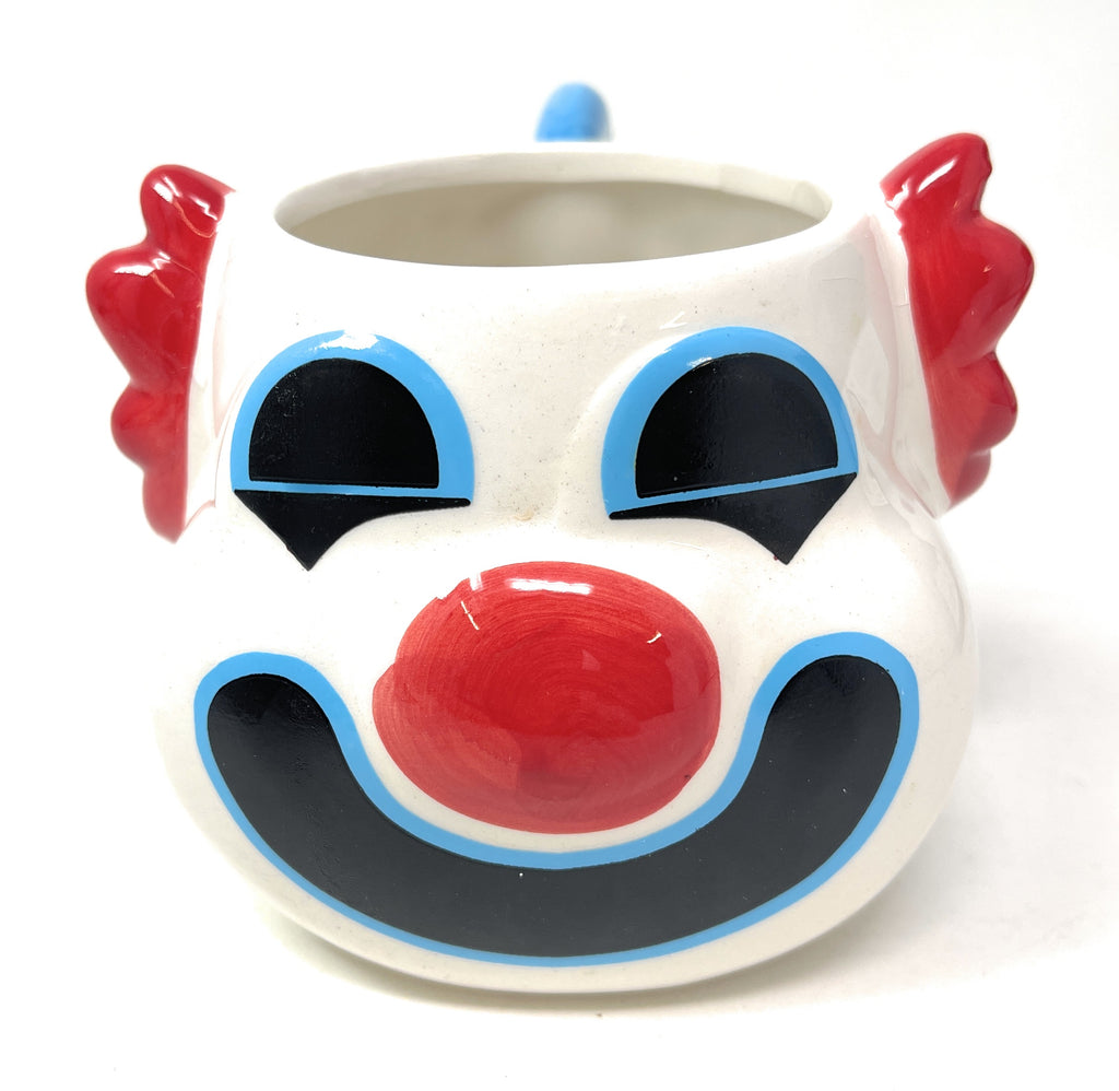 A white ceramic clown mug shaped like a clown face, featuring a glossy red nose, red hair on the sides, and blue and black painted details on a white background.