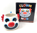 A novelty clown face heat-changing mug shown next to its black retail box. The mug is white with a red nose, red hair, blue outlines, and black facial features.
