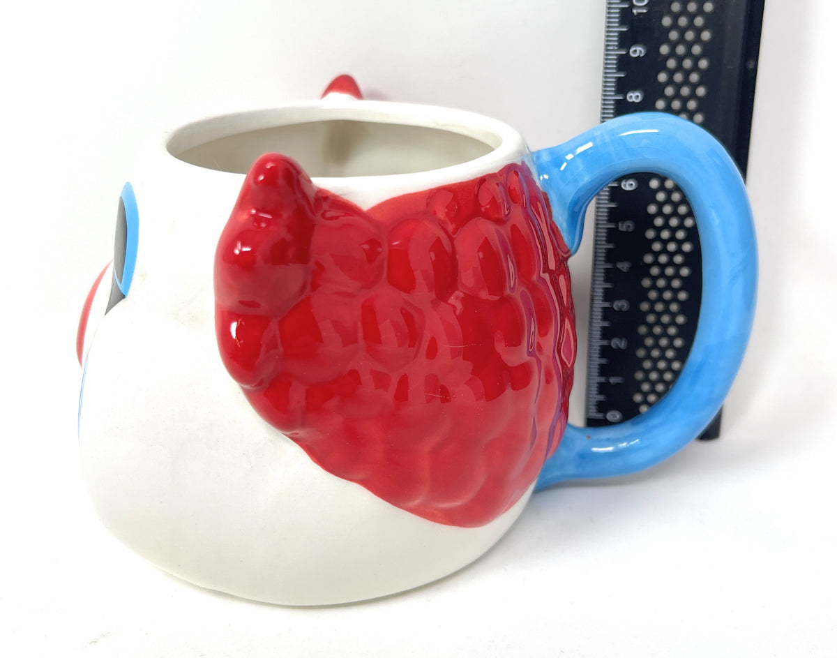 A white ceramic mug  clown face, featuring a large red 3D  ear on the side and a bright blue handle. A black ruler is positioned behind the mug to show its scale.